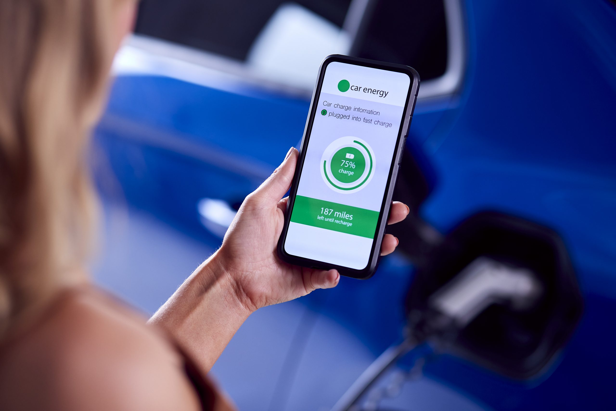 Woman Charging Electric Car With Cable Using App On Phone To Monitor Battery Level