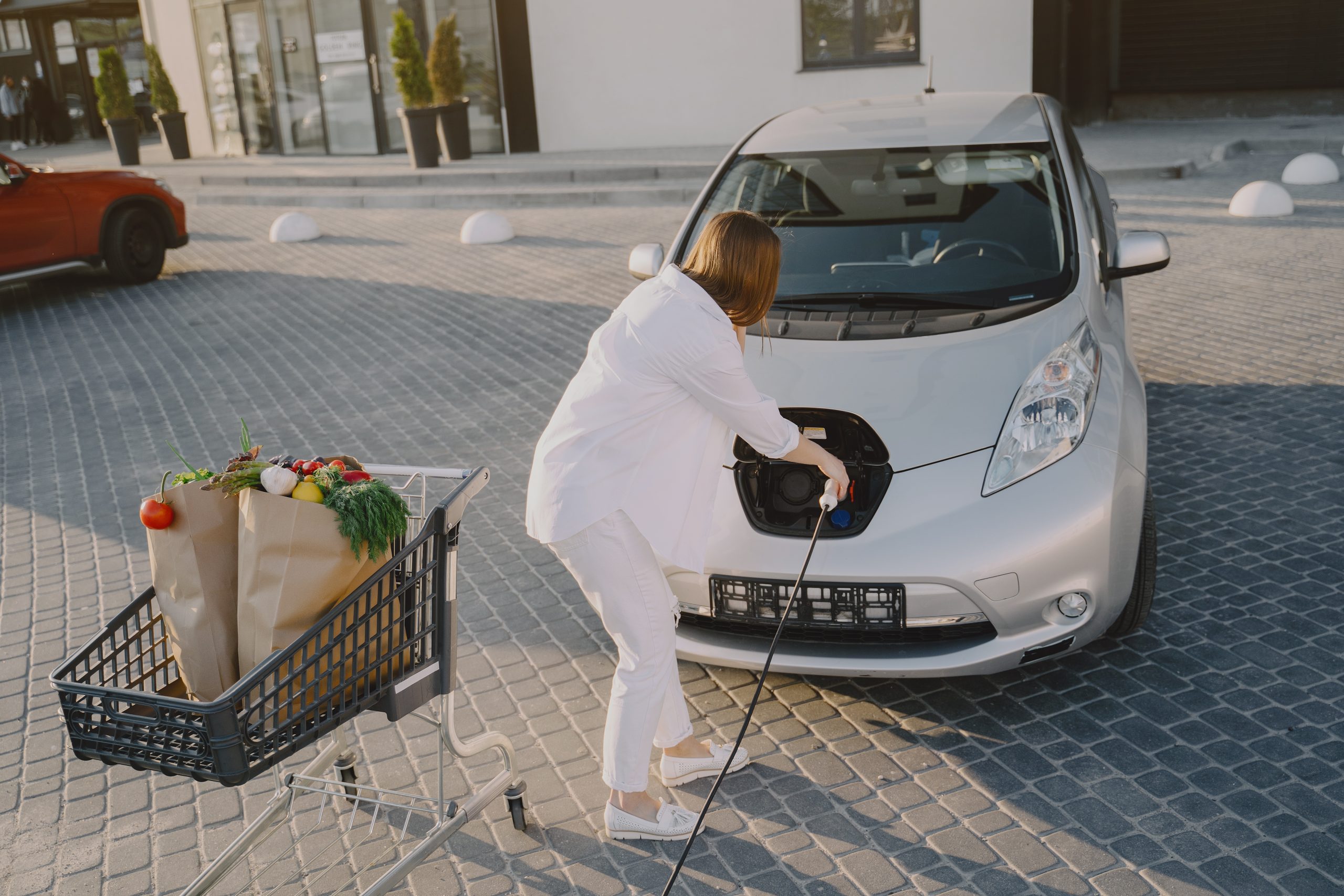 Charging electro car at the electric gas station. Woman standing by the car. Lady with foodstuff.